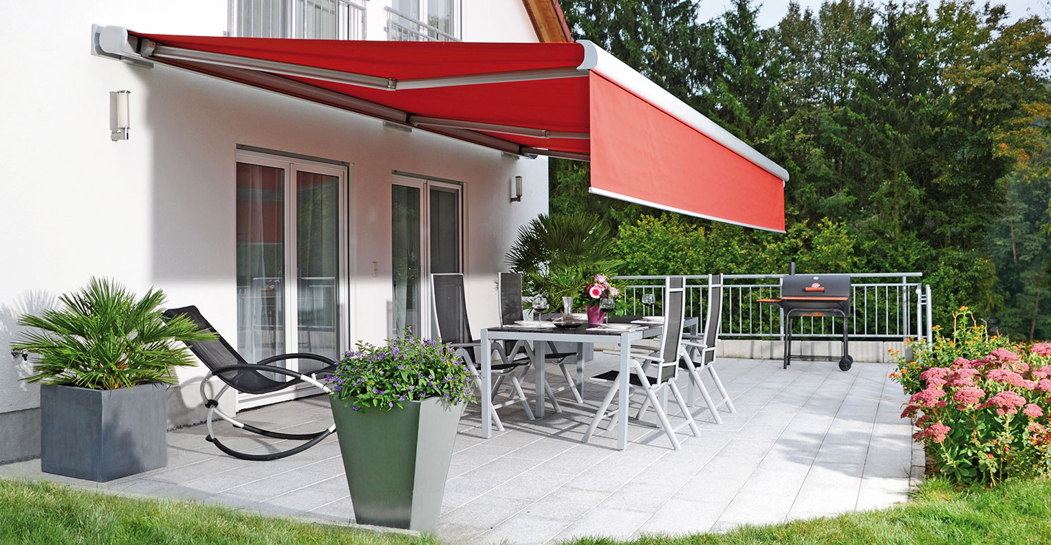 Rote Markise an der Hausfassade über einer Terrasse mit Garten