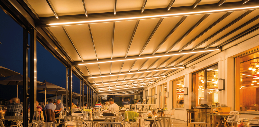 Pergola mit geschlossenem Faltdach, LED-Beleuchtung an einer Gastronomie mit Sitzgelegenheiten und vereinzelt Personen